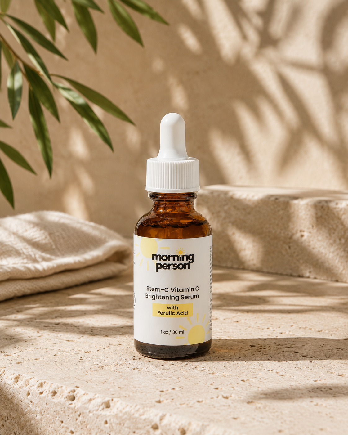 A 1 oz amber glass dropper bottle of Morning Person Stem-C Vitamin C Brightening Serum with Folic Acid, set on a textured beige stone surface with soft shadows and green leaves in the background.