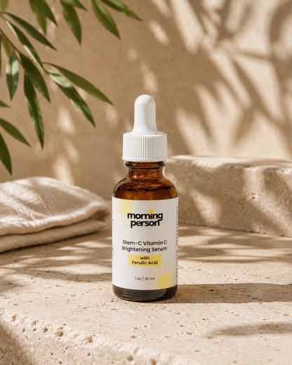 A 1 oz amber glass dropper bottle of Morning Person Stem-C Vitamin C Brightening Serum with Folic Acid, set on a textured beige stone surface with soft shadows and green leaves in the background.