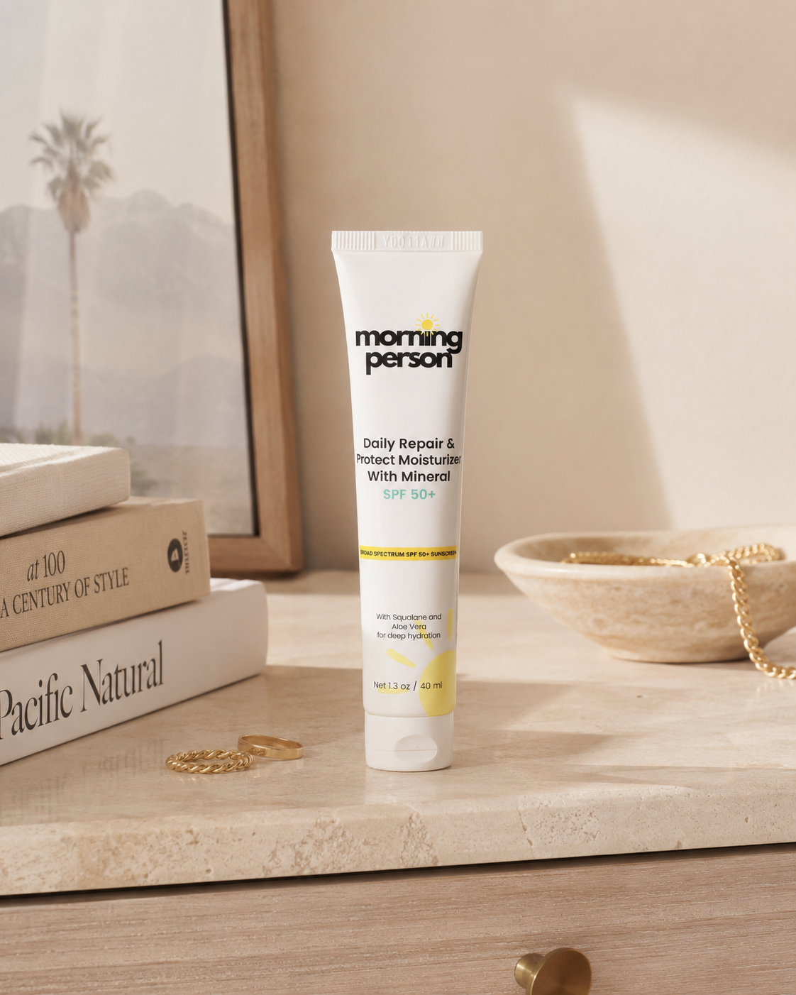 A white tube of Morning Person Daily Repair &amp; Protect Moisturizer With Mineral SPF 30 stands on a light stone surface in a styled interior scene with books, jewelry, and a bowl.