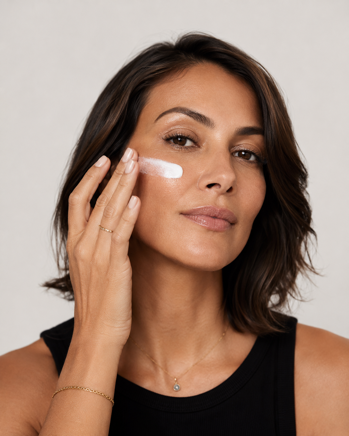 A woman with dark brown hair applies a non-nano mineral spf 50 product to her cheek against a light gray background.