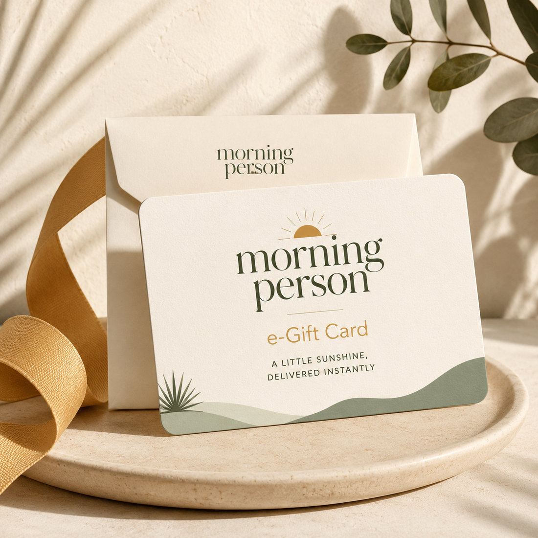 A Morning Person e-Gift Card with its matching envelope, styled on a beige ceramic tray with a gold ribbon and soft leaf shadows.